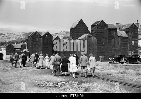 Holiday scenes. in and around Hastings, East Sussex.. Tourist walk along the seafront past the wooden sheds that holding the fishing nets of the local fishermen. June 1952. Stock Photo