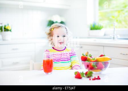 Beautiful toddler girl with curly hair wearing a colorful shirt having breakfast drinking juice in a white sunny kitchen Stock Photo