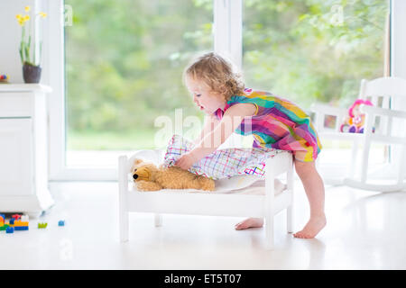 Sweet curly toddler girl playing with her teddy bear putting him in toy bed to sleep in sunny room with big garden view windows Stock Photo
