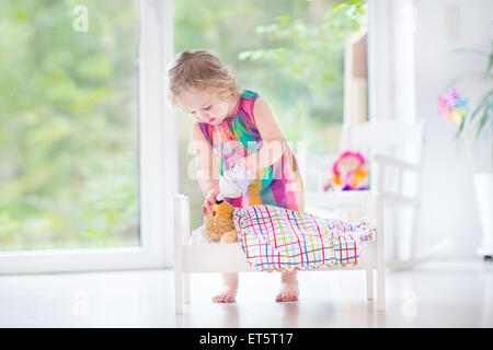 Sweet curly toddler girl playing with her teddy bear putting him in toy bed to sleep in sunny room with big garden view windows Stock Photo