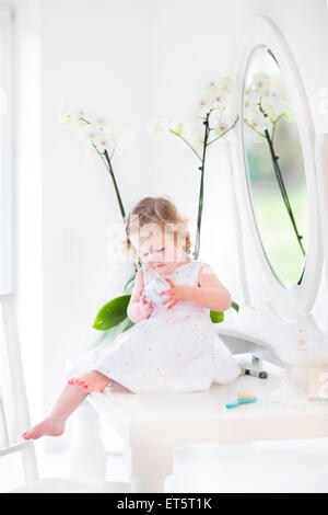 Happy toddler girl with curly hair wearing a white dress playing with make up and cosmetics in front of a round mirror Stock Photo