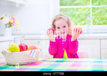 Funny happy laughing child, adorable toddler girl with curly hair wearing a pink shirt, eating red and green apples for snack Stock Photo