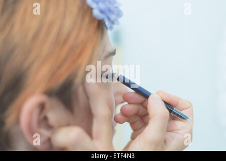 Mature woman applying eyeliner on eyelid with blue pencil. Neutral background, toned image, decontrasted. Selective focus. Stock Photo