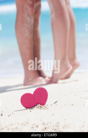 Barefoot female legs standing up tiptoe on man's foots on beach with turquoise sea background, decorated pink heart object.  Rom Stock Photo