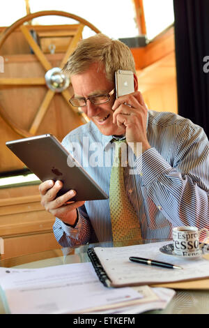 Online business meeting home alone businessman houseboat office at table with notes looking at iPad air tablet computer screen using iPhone smartphone Stock Photo