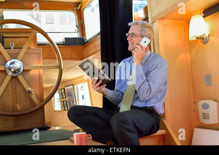 Relaxed mature businessman in houseboat office holding his iPad air tablet computer screen and using his iPhone 6 smartphone Stock Photo