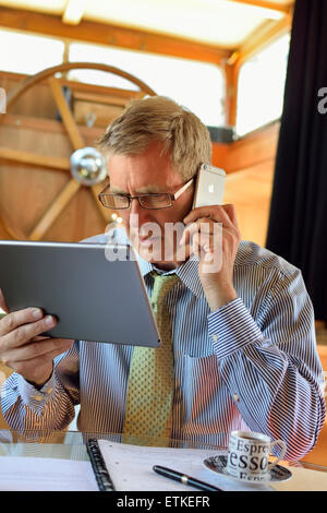Man mature 50s checking finances working in houseboat office using iPad tablet & smartphone concentrates on the latest serious screen information Stock Photo