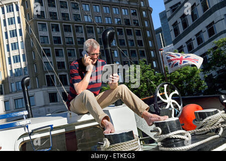 CANARY WHARF mature businessman sitting outside on his office boat talking on smartphone looking at his iPad, offices behind Canary Wharf London E14 Stock Photo