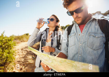 Couple on country walk together, summer vacation in countryside. Young man reading a map while woman drinking water in backgroun