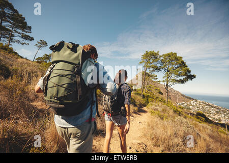 Rear view of two young people walking down the trail path on mountain. Young couple hiking with backpacks. Stock Photo
