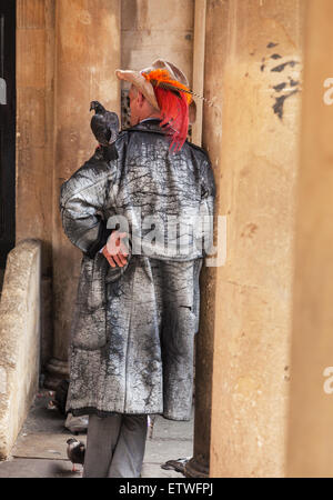 Man leaning against a stone pillar with a pigeon on his shoulder, wearing a feathered hat. Stock Photo