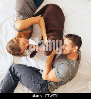 Top view of happy young family relaxing on bed together. Man and woman with pet dog in bedroom. Stock Photo