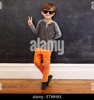 Full length shot of cool little boy wearing sunglasses gesturing victory sign. Young school boy in casuals showing peace symbol Stock Photo