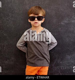 Portrait of innocent little kid wearing sunglasses. Little boy leaning on a blackboard. Square composition. Stock Photo
