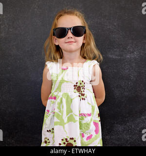 Portrait of beautiful little girl wearing sunglasses posing at camera against a black wall. Stock Photo