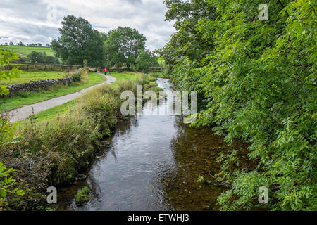 A small river running through the countryside with pedestrians walking along a footpath to the left of the river Stock Photo