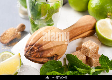 Wooden juicer and ingredients for making mojitos. Ice, fresh mint leaves sugar cubs, lime and glass on white cutting board Stock Photo