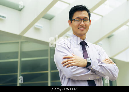 Portrait of happy and confident young asian businessman. The man stands in a street against office buildings and crosses arms lo Stock Photo