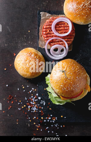 Fresh homemade burger on black slate and raw cutlet and sliced onion, served with sea salt and pepper over dark background. Sele Stock Photo