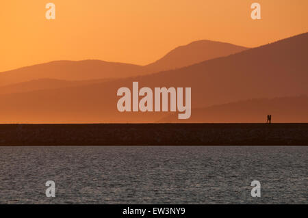 Silhouetted walkers on a jetty at the moment of a golden sunset, with sea in foreground and mountains in background Stock Photo