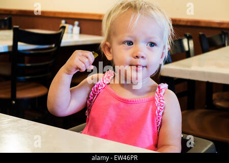 An 18 month old blonde Caucasian baby girl enjoys a sour pickle at a restaurant. USA. Stock Photo