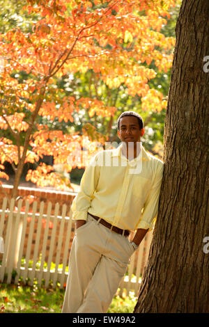 A young hispanic male leans against an old tree trunk in the early fall with orange leaves behind. Stock Photo