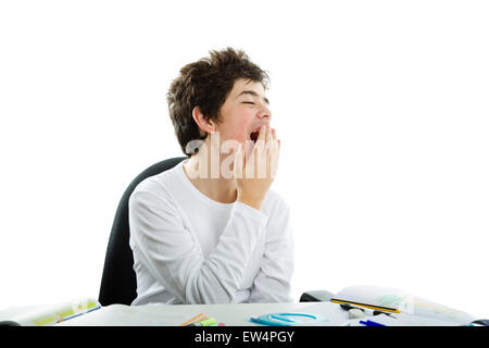 Tired Caucasian boy sits in front of homework wearing a white long sleeve t-shirt and yawns covering his mouth with right hand Stock Photo
