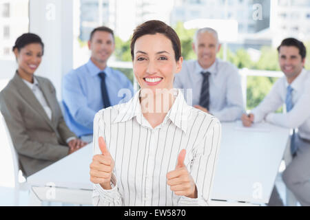 Smiling businesswoman giving thumbs up with her team behind Stock Photo