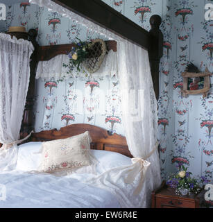 White lace drapes on four poster bed in nineties cottage bedroom with patterned wallpaper Stock Photo