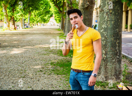 Funny attractive young man standing, wearing yellow T-shirt and blue jeans while eating a tasty vanilla ice cream outdoors in th Stock Photo