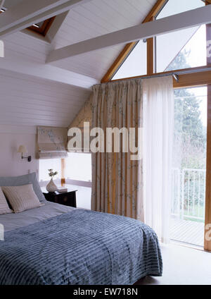 Blue-gray quilt on bed in modern loft conversion bedroom with floral drapes and white voile curtains at glass doors to balcony Stock Photo
