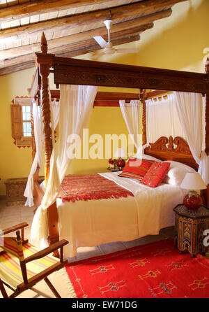 Red cushions and ethnic rug on rustic wooden four poster bed with white drapes in yellow Spanish country bedroom Stock Photo