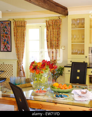 Vase of orange tulips on a glass topped table in yellow cottage dining room with patterned curtains on window Stock Photo