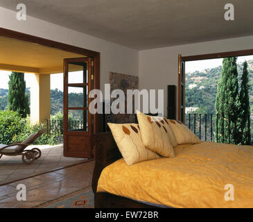 Cream cushions and yellow bed cover on bed in bedroom in modern Spanish villa with doors open to the terrace Stock Photo