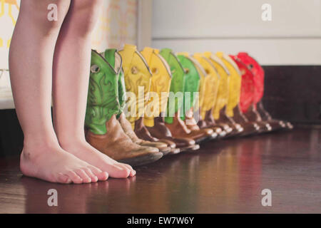 Barefoot boy standing next to a row of multi coloured cowboy boots Stock Photo