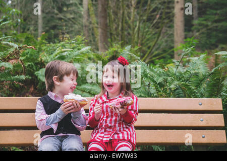 Two children sitting on a bench laughing and eating donuts Stock Photo