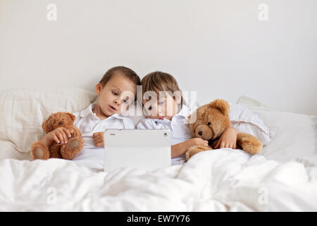 Two cute boys, sitting in bed, playing with tablet, teddy bears in their hands Stock Photo