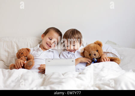 Two cute boys, sitting in bed, playing with tablet, teddy bears in their hands Stock Photo