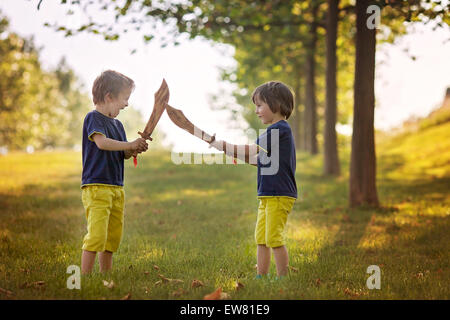 Two little boys, holding swords, glaring with a mad face at each other, fighting outdoors in the park Stock Photo