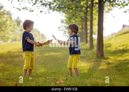 Two little boys, holding swords, glaring with a mad face at each other, fighting outdoors in the park Stock Photo