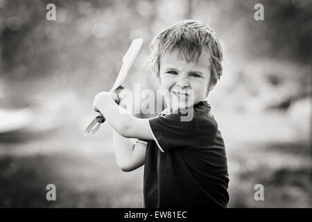 Angry little boy, holding sword, glaring with a mad face at the camera, outdoors in the park, monochrome conversion Stock Photo