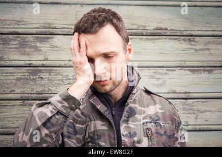 Young Caucasian man with headache in camouflage. Outdoor portrait over green rural wooden wall Stock Photo