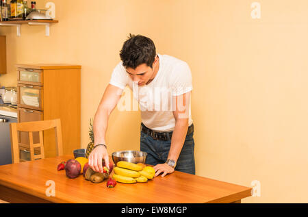 Young man preparing a healthy fruit salad or smoothie, standing leaning on a kitchen counter loaded with an assortment of fresh Stock Photo