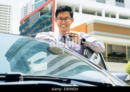 Young adult chinese businessman holds keys of new car, leaning on auto door. He smiles and looks proud at camera, showing the ke Stock Photo