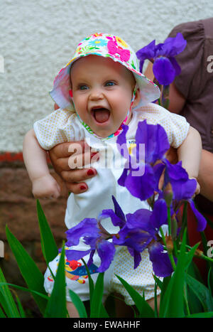 The five-month girl cheerfully laughs, close to the blue irises Stock Photo