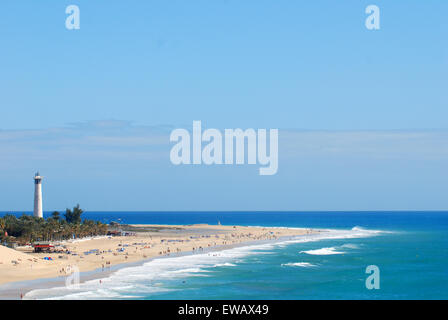 A sunny sand beach being hit gently by foamy wave crests, with a lighthouse standing on a pouch of tropical trees. Stock Photo