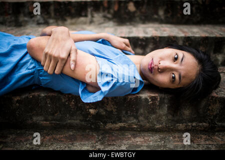 A young Vietnamese woman in a blue dress, lying on stone steps in rural Hanoi, Vietnam. Stock Photo
