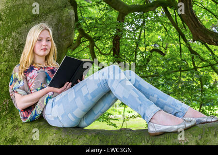 Blonde caucasian teenage girl lying on green tree reading book Stock Photo