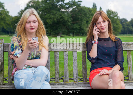 Two caucasian teenage girlfriends sitting on wooden bench in park calling with mobile phone Stock Photo