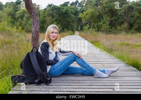 Blonde caucasian teenage girl resting on wooden footpath in forest area Stock Photo